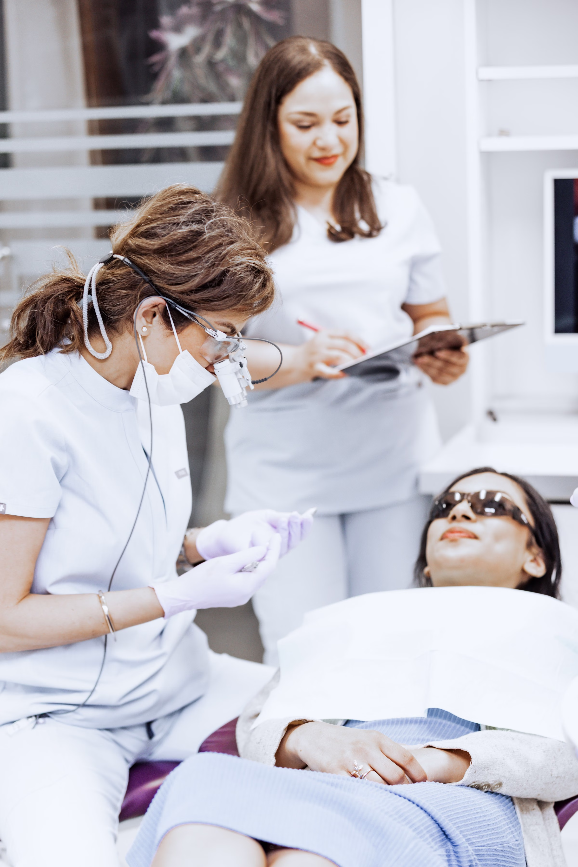 A cosmetic dentist wearing loupes and a mask examines a patient in the modern treatment room at Georgetown Cosmetic Dentistry in Washington DC while an assistant takes notes on a clipboard.
